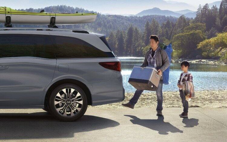 2026 Honda Odyssey Elite parked at lake with hills and greenery in background. Father using the motion sensor to open the back door to load items while son is watching.